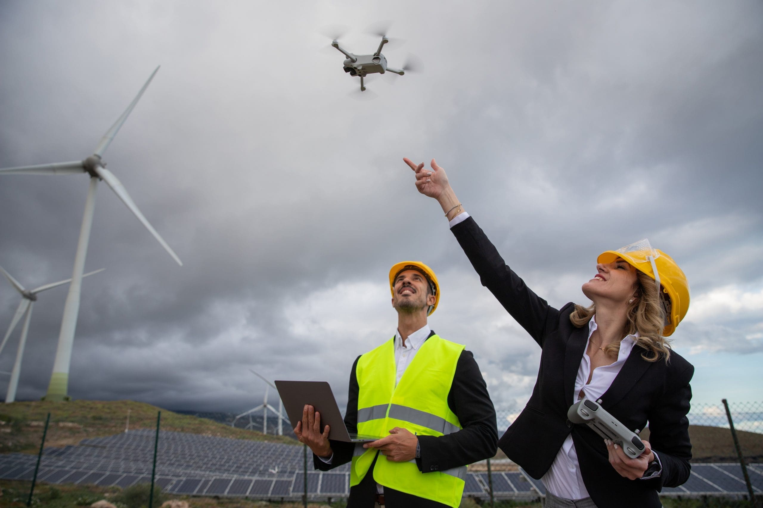 Two engineers at work fly a drone and use the laptop in a solar farm with wind turbines.